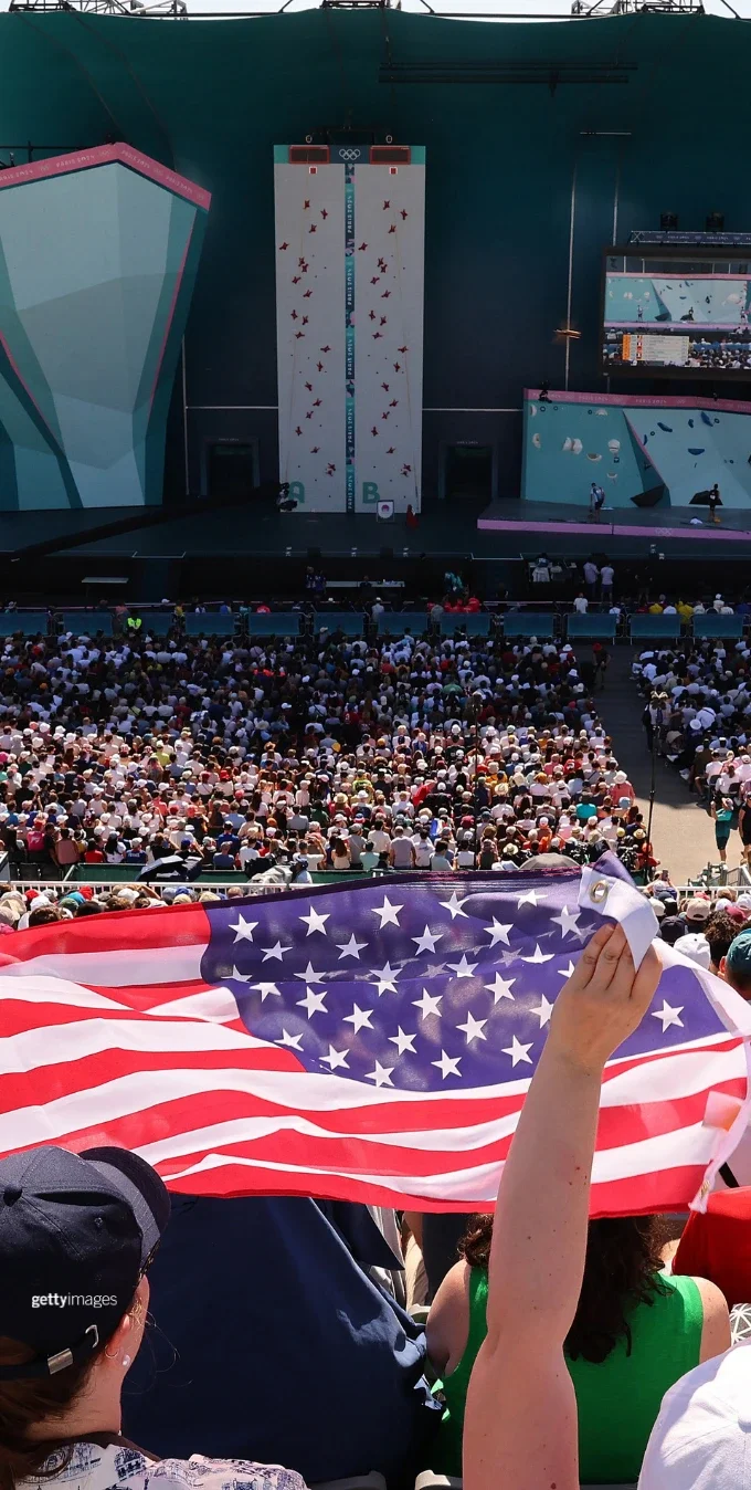 A photo of fans cheering on a sport climbing competition at the Paris 2024 Summer Games. The foreground has a fan waving the American flag.