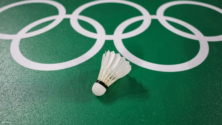 Un volante de bádminton descansa sobre una cancha verde con el logo de los anillos olímpicos blancos al fondo. La textura del suelo de la cancha aporta detalles a la imagen en primer plano.