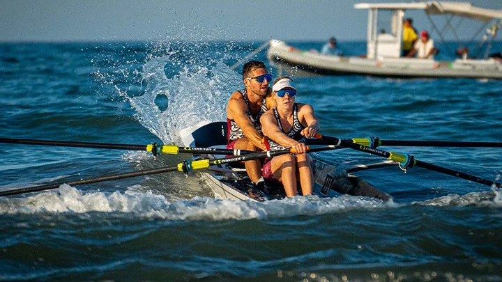 A photograph of a mixed team of a female rower in the front and a male rower in the back out on the ocean with a white boat with several people on board in the background.