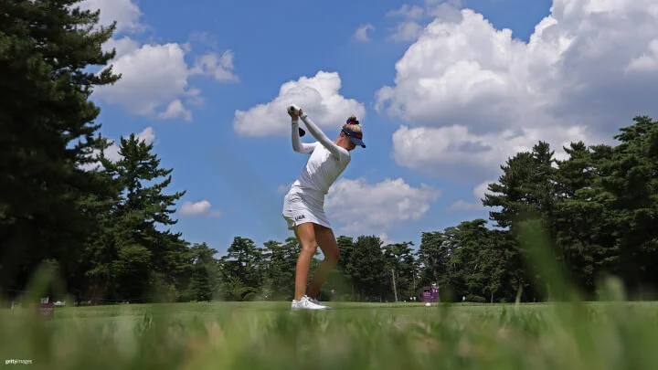 Una golfista femenina es capturada en medio de su swing en un día brillante y soleado. Ella lleva puesta una camisa blanca de manga larga, una falda a juego y una visera azul oscuro con las letras "USA" visibles en su vestimenta. El fondo muestra un cielo azul claro con nubes blancas dispersas y árboles verdes rodeando la escena. La perspectiva está cerca del suelo, con césped verde en primer plano.