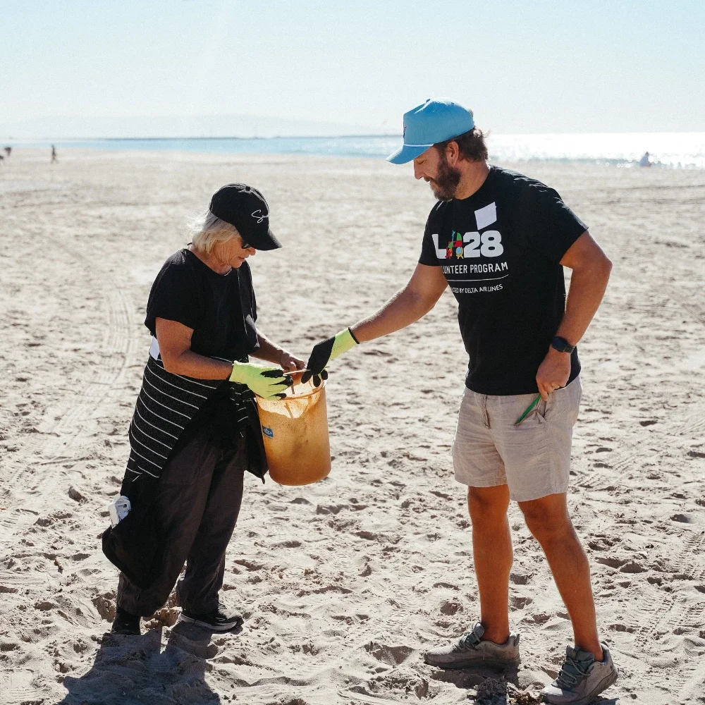 Woman holding a bucket next to a man in a baseball cap with the sand in the forefront and ocean in the background