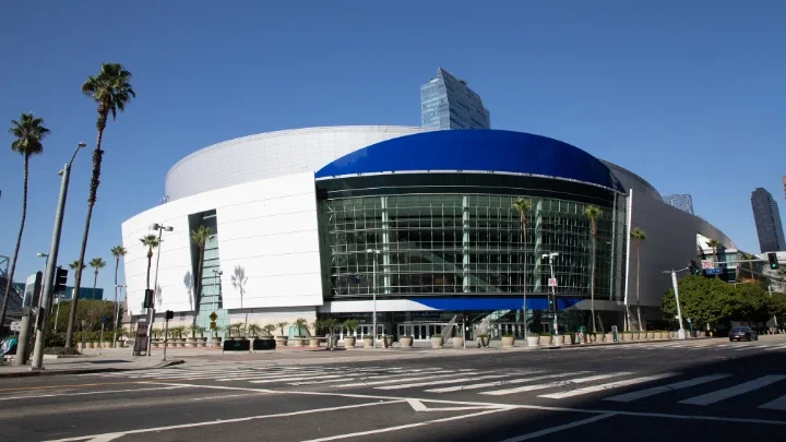 A photograph of the Arena in Downtown Los Angeles (Crypto.com Arena) showing the front entrance with a curved building design and glass windows. Palm trees are shown on the left side of the photograph by the sidewalk.