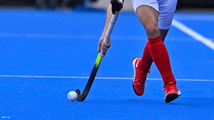 A close-up image of a field hockey player in motion on a bright blue turf field. The player is wearing red socks, red shoes, and white shorts. They are holding a black and neon yellow field hockey stick, controlling a white hockey ball on the ground. The focus is on the player's lower body and the stick.