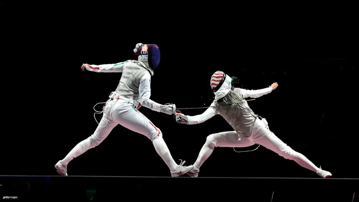 Two fencers in full gear are competing on a dark background. The fencer on the left, wearing a blue mask, lunges forward with their sword. The fencer on the right, wearing a red and white striped mask, also lunges to counter the attack. Both athletes are in white uniforms with silver vests.