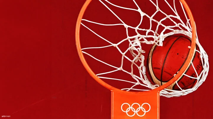 A basketball is seen going through a hoop, captured at the moment it is through the net. The hoop and net are standard in design, with the rim being orange and the net white. The backboard to which the hoop is attached features the Olympic rings logo, indicating that this is an Olympic basketball event. The background is a solid red color.