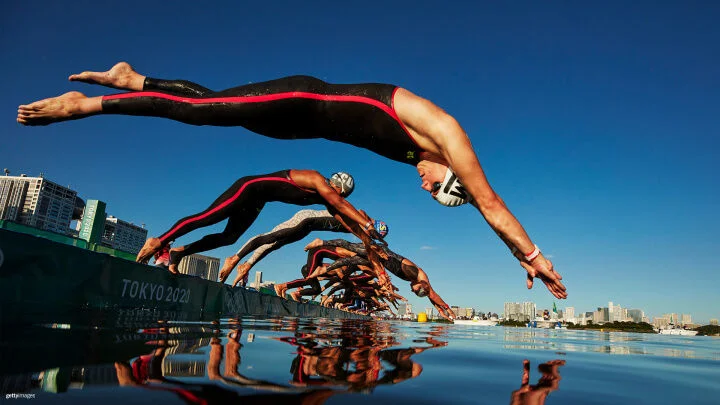 A group of swimmers are captured mid-dive at the start of the swimming portion of the Tokyo 2020 Olympics. The athletes, wearing black wetsuits with red stripes on the side and swim caps, are in perfect alignment as they dive into the water against a clear blue sky. The city's skyline and Olympic signage are visible in the background.