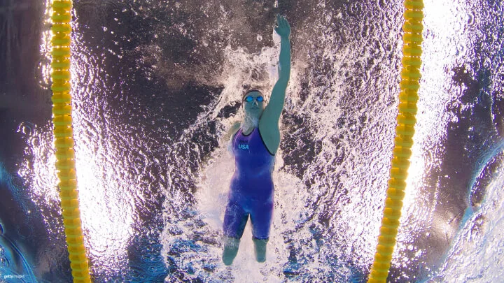 An underwater shot captures a female Paralympic swimmer from Team USA during a freestyle event. She is wearing a blue swimsuit, swim cap, and goggles. Her left arm is extended upward as she glides through the water, creating ripples and bubbles around her. The swimmer's legs are amputated below the knee, and her right arm is also amputated. Yellow lane dividers are visible on both sides, framing her path in the pool.