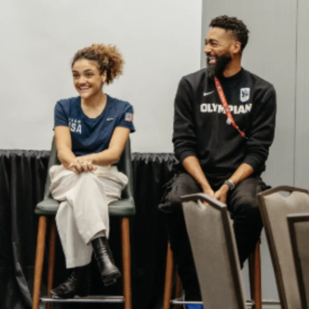 A photograph of Olympic gymnast Laurie Hernandez wearing a navy blue Team USA shirt sitting next to Jason Richardson who was wearing a black sweater with the word Olympian on it, on stools during a panel talk