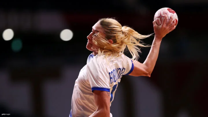 Una toma media captura a una jugadora femenina de balonmano, con cabello rubio recogido en una cola, en acción a punto de lanzar el balón. Ella lleva puesta una camiseta blanca con detalles azules. El nombre en la parte posterior de su camiseta no es completamente visible.