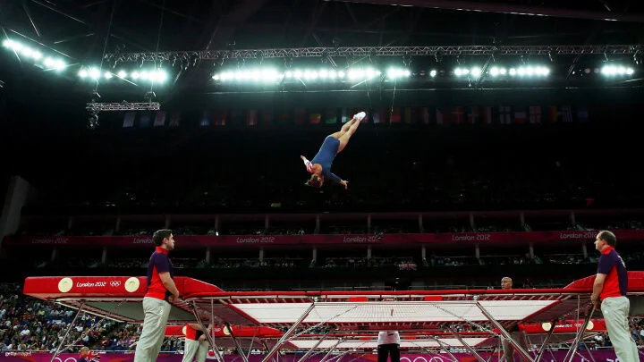 A gymnast in a blue leotard performs a high-flying routine on a trampoline. She is captured mid-air, fully extended with her body parallel to the ground and her arms outstretched to the side. The trampoline is surrounded by spotters in red and purple uniforms. The background shows a large arena filled with spectators, Olympic banners, and bright overhead lights. National flags from various countries hang above the crowd.