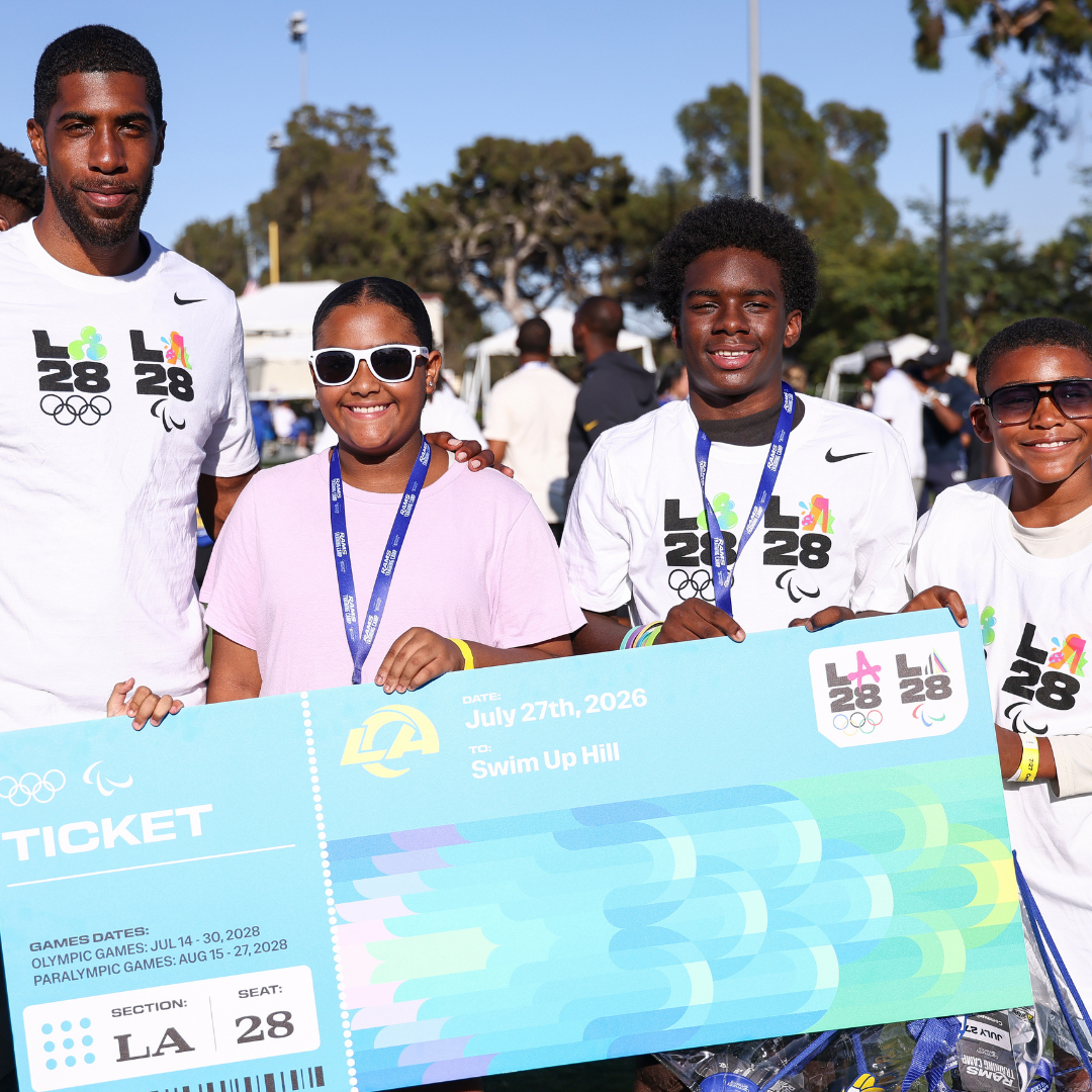 Team USA Paralympian Jamal Hill wearing a white t-shirt standing with children in white LA28 t-shirts while holding a large blue ticket poster. 