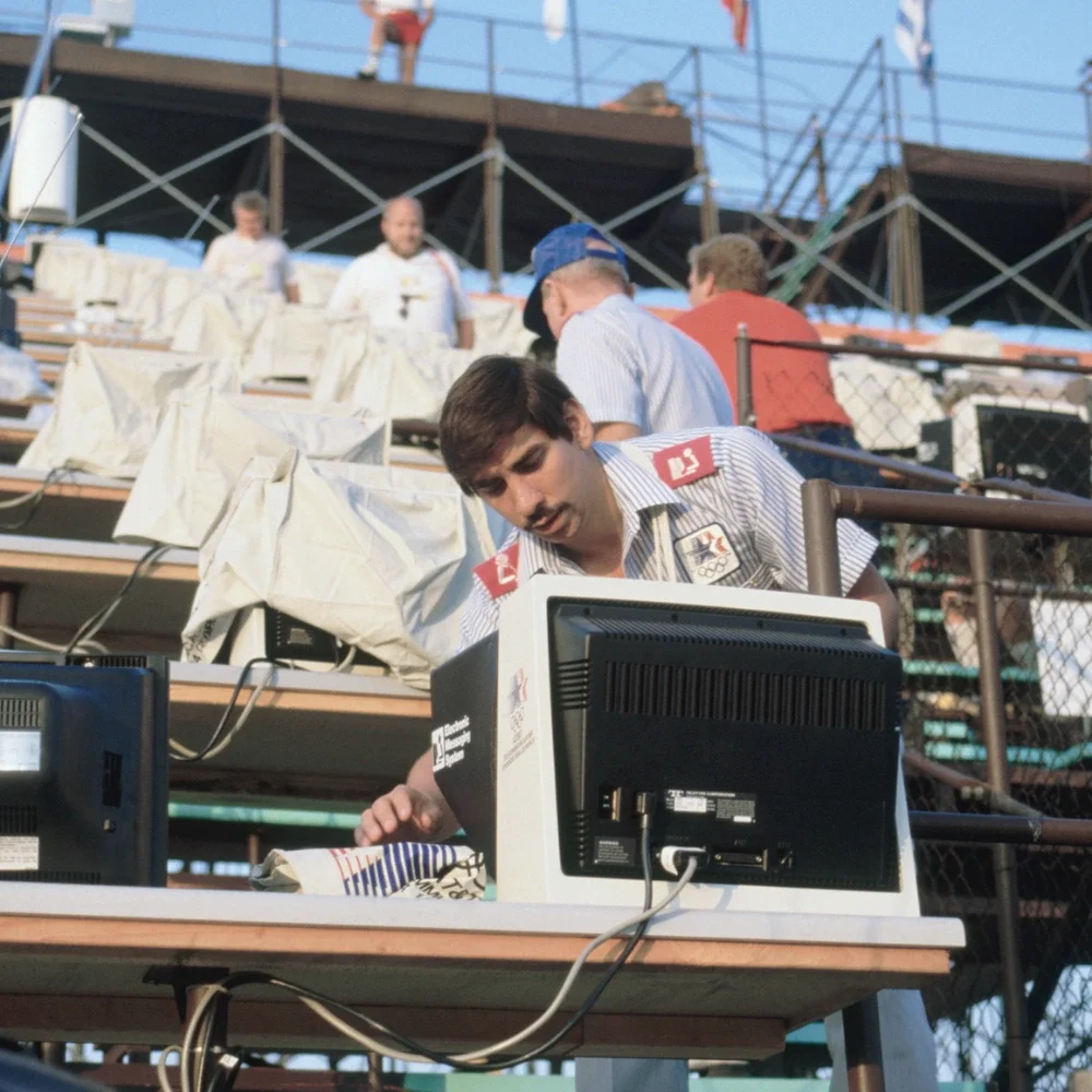 Photograph of man fixing a computer for the electronic messaging system during the 1984 Games, rows of computers and other people working on the computers are in the background at an outdoor venue