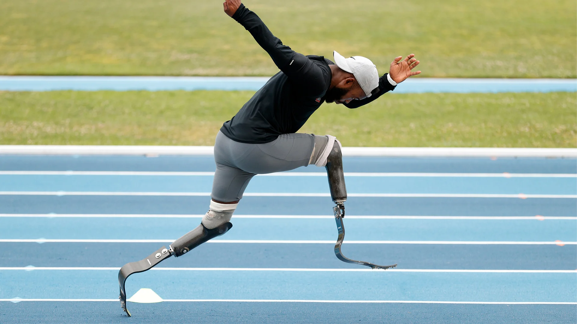 A photo of an African American male with prosthetic legs wearing grey shorts and a long sleeved black shirt and a white hat sprinting on a blue track