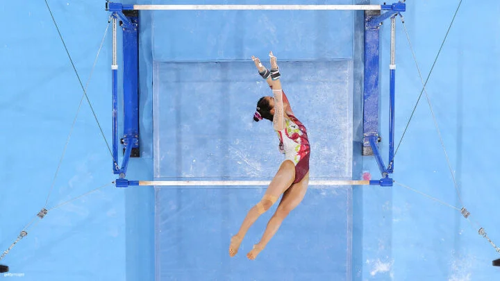 Una gimnasta femenina es capturada en el aire durante su rutina en las barras asimétricas. Se le ve desde arriba, saltando entre las barras con el cuerpo de lado, los brazos extendidos y los dedos apuntando. Lleva puesto un maillot rojo, verde y blanco con diseños intrincados. Su cabello está recogido en un moño y sus muñecas están envueltas con cinta protectora y agarraderas. El fondo muestra un colchón azul de gimnasia con marcas de tiza.