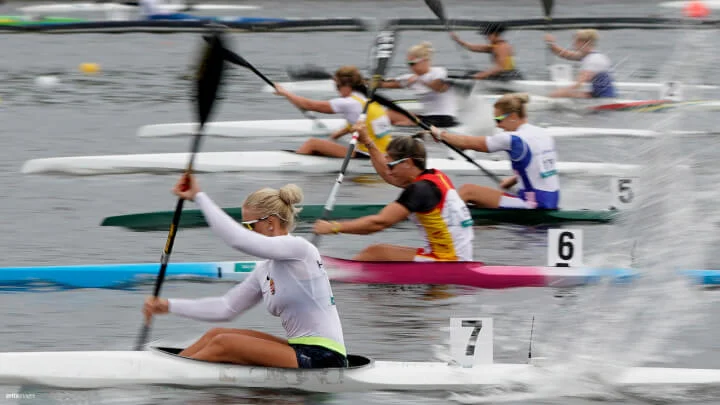 A dynamic shot of a canoe race on a calm body of water, featuring multiple female athletes paddling vigorously. The canoes are in close proximity, with visible lane markers numbered from right to left, 1 to 7. The athlete in the foreground, wearing a white long sleeve shirt and sunglasses, is in kayak number 7. The competitors are captured in mid-stroke, creating splashes as their paddles hit the water.