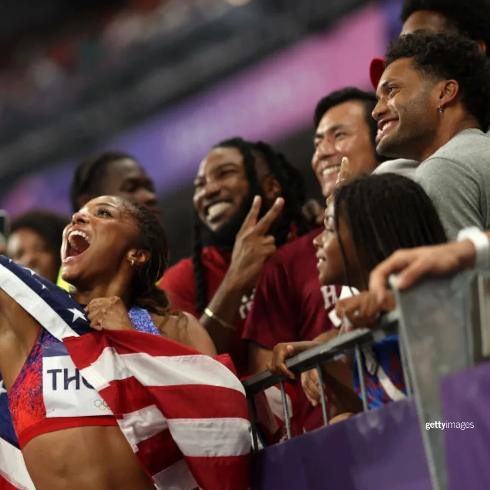 A photograph of a female track and field athlete smiling and taking a photo with fans in the stands