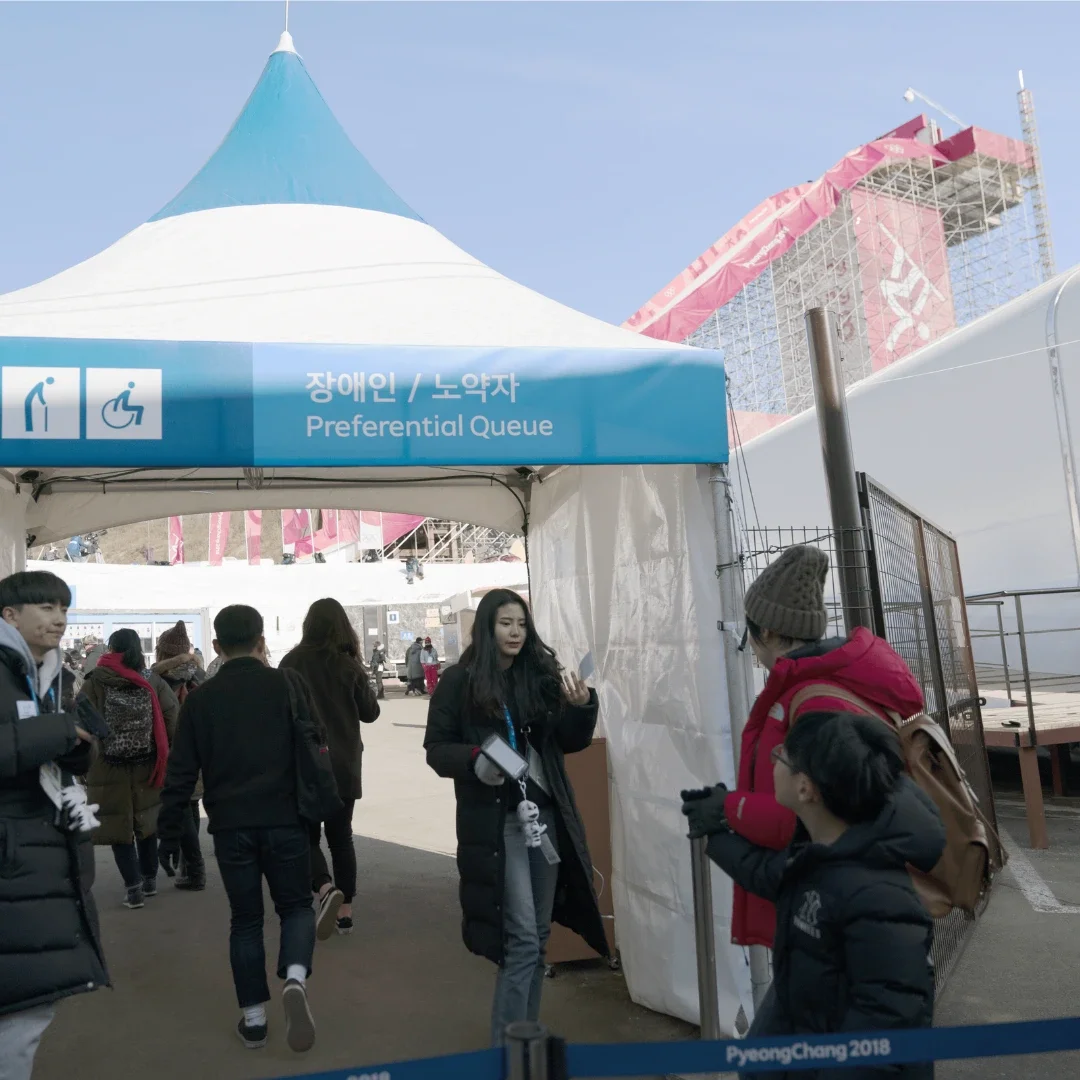 Fans walking into an Accessibility entry checkpoint while a woman is looking at her phone to check them in. A tent is in the background with a blue trim with images of different abilities including a pregnant woman pictogram, a man on crutches, a man bending over with a cane and a person in a wheelchair. 