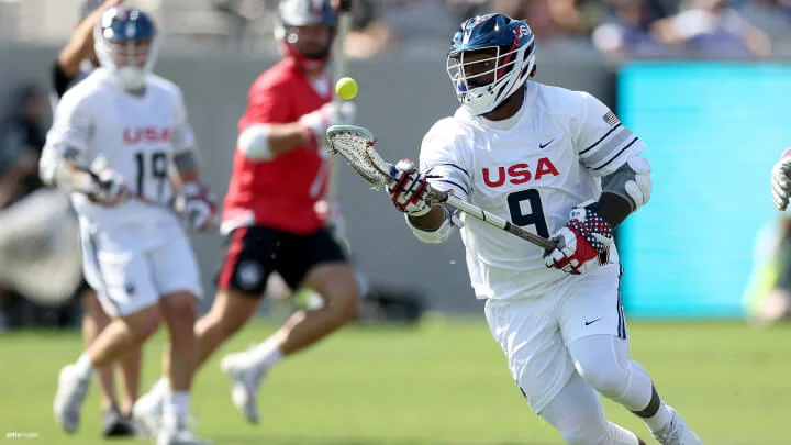 Un jugador de lacrosse masculino, vistiendo un uniforme blanco con "USA" y el número 9 en el frente, está en acción en el campo. El jugador sostiene un palo de lacrosse y parece estar siguiendo o a punto de atrapar una pelota amarilla de lacrosse en el aire. Adicionalmente, el jugador tiene puesto un casco blanco con visera azul, guantes blancos y protectores de brazo. En el fondo, se distingue a otro jugador con uniforme blanco y a uno más con uniforme rojo, mientras el juego se desarrolla en un campo verde.