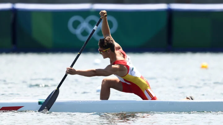 A male canoeist, dressed in a red, yellow, and white athletic uniform, powers through calm water during an Olympic event. The athlete is in a striding position with one knee on the deck and the other foot forward. He has his paddle submerged in the water as he propels his white canoe forward. The background is blurred and features Olympic branding.