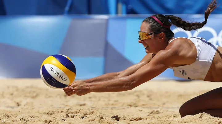 Una jugadora de voleibol de playa usando un sujetador deportivo blanco y gafas de sol, se lanza a la arena para realizar una defensa. Tiene una expresión concentrada y su cabello está recogido en una cola. El balón  es amarillo, azul y blanco, marcado con "Tokio 2020". El fondo muestra una vista borrosa de los anillos olímpicos y una barrera azul.