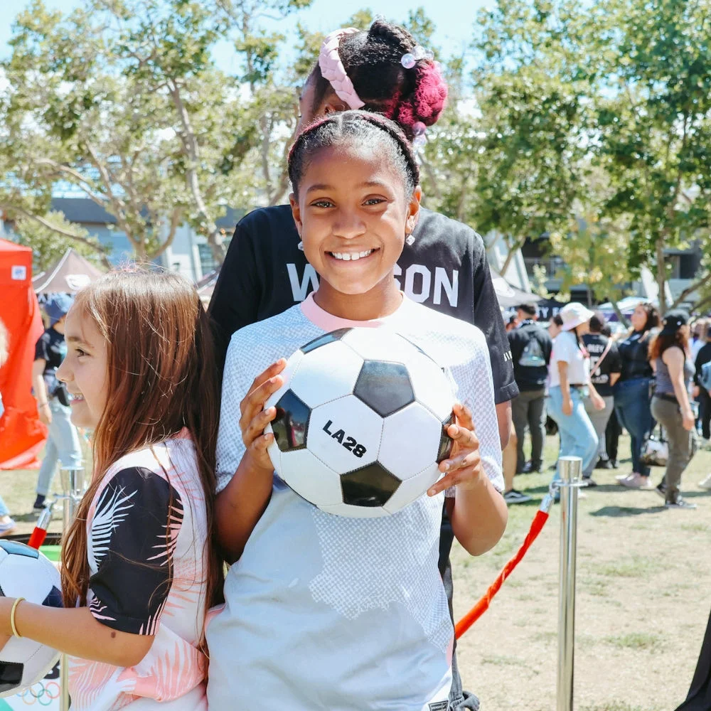 Foto de una niña con camiseta blanca sosteniendo un balón de fútbol con LA28 imprimido en el