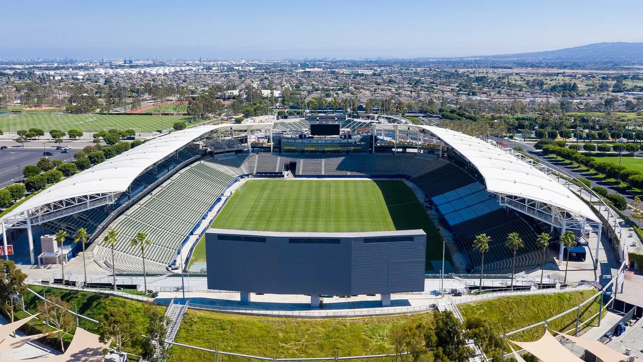 An aerial photograph of Dignity Health Stadium showing empty stands on three sides and a large empty green field of grass. The Stadium is surrounded by grass and trees. In the background the city scape is visible featuring many buildings and homes as well as a large hill on the right and blue sky.