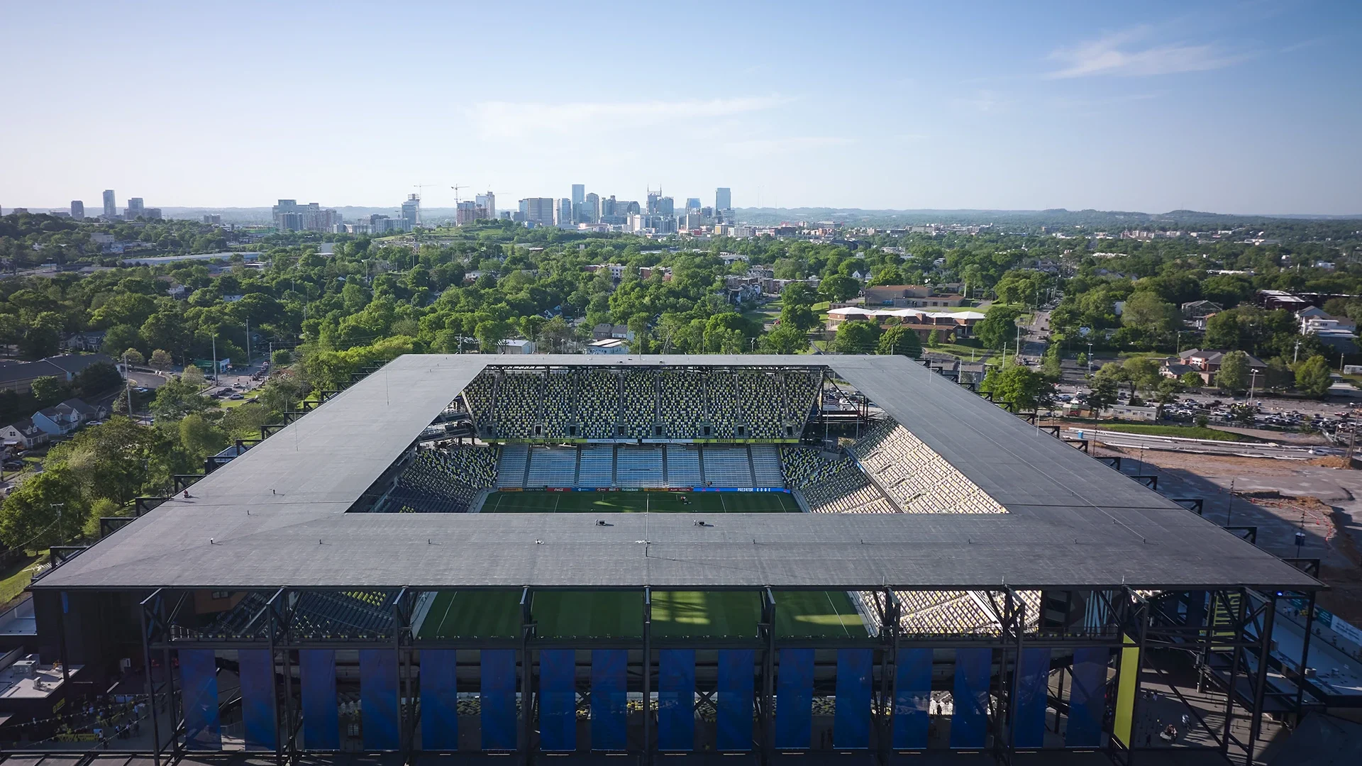 Aerial view of Nashville Stadium with an open-air roof and fan seating in the middle of the stadium. Trees and houses and the downtown buildings in Nashville are shown in the background.