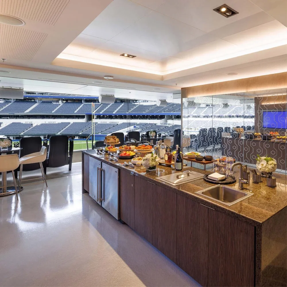 A hospitality suite at Sofi Stadium with a food setup inside the suite. An island with a sink and dark wood cabinetry is shown in the foreground.