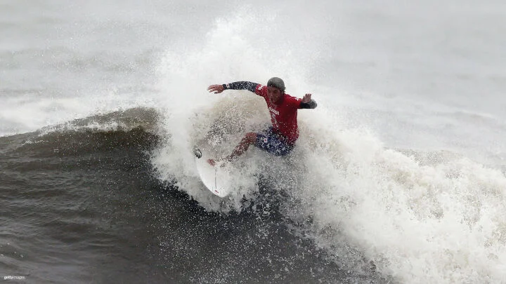 Un surfista con una camiseta roja y azul y pantalones cortos azules surfea una ola grande. El cuerpo del surfista está posicionado de lado, con los brazos extendidos para mantener el equilibrio, mientras las espumas blancas salpican a su alrededor.