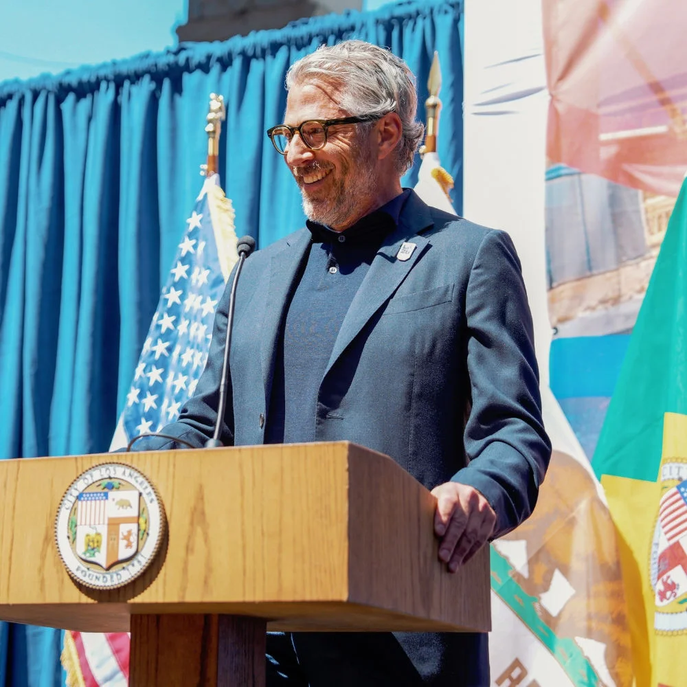 Fotografía mostrando el presidente de LA28, Casey Wasserman, de pie en un podio con un micrófono durante la ceremonia de instalación de la bandera olímpica y paralímpica en el Ayuntamiento de Los Ángeles.