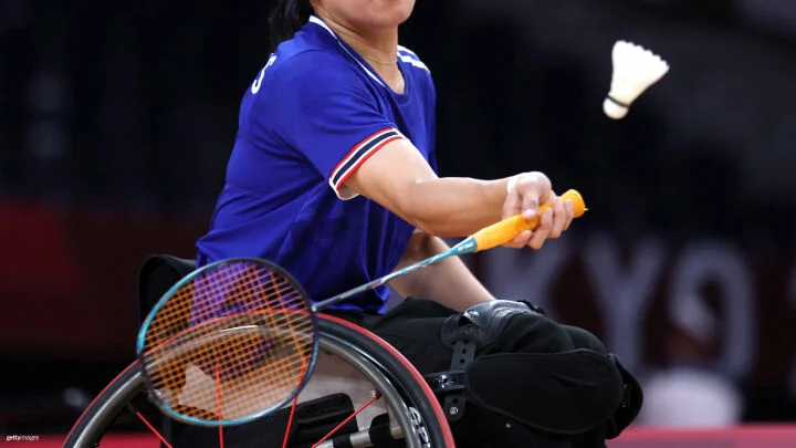 Una imagen cercana de un jugador de bádminton en silla de ruedas golpeando un volante con una raqueta. El jugador lleva una camiseta azul con rayas rojas y blancas en las mangas, y sostiene una raqueta con mango de color naranja. La atención se centra en el brazo del jugador y la raqueta a punto de golpear el volante que está en el aire.