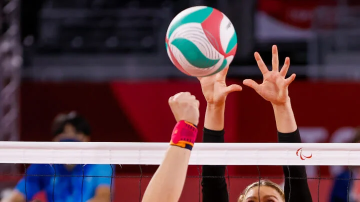 Two sitting volleyball players are reaching up towards a multicolored volleyball above the net. The player on the left, wearing a pink and orange wrist brace, extends their closed fist upwards. The player on the right has both hands open with fingers spread wide, preparing to block the ball. The background is blurred, with a red color scheme.