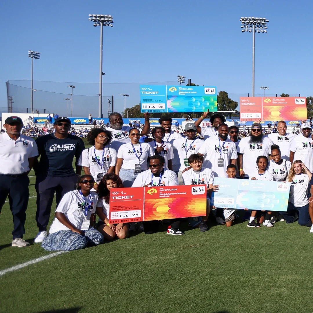 Group of people holding up large LA28 ticket recipient posters while standing on a grass football field.