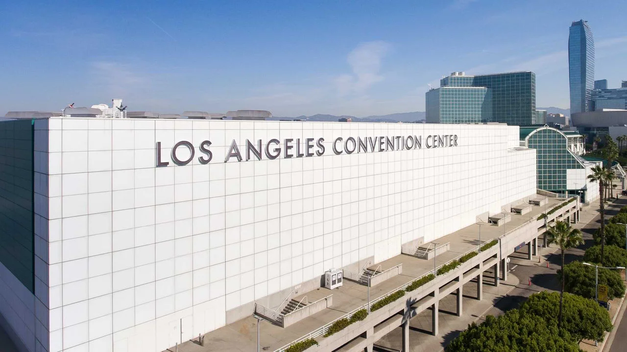 Fotografía mostrando el Los Angeles Convention Center en el centro de Los Ángeles. Es un gran edificio rectangular blanco con las palabras "Los Angeles Convention Center" en letras grandes plateadas. El diseño del edificio tiene varios cuadros pequeños que cubren toda la estructura con el perfil urbano de Los Ángeles en el fondo.