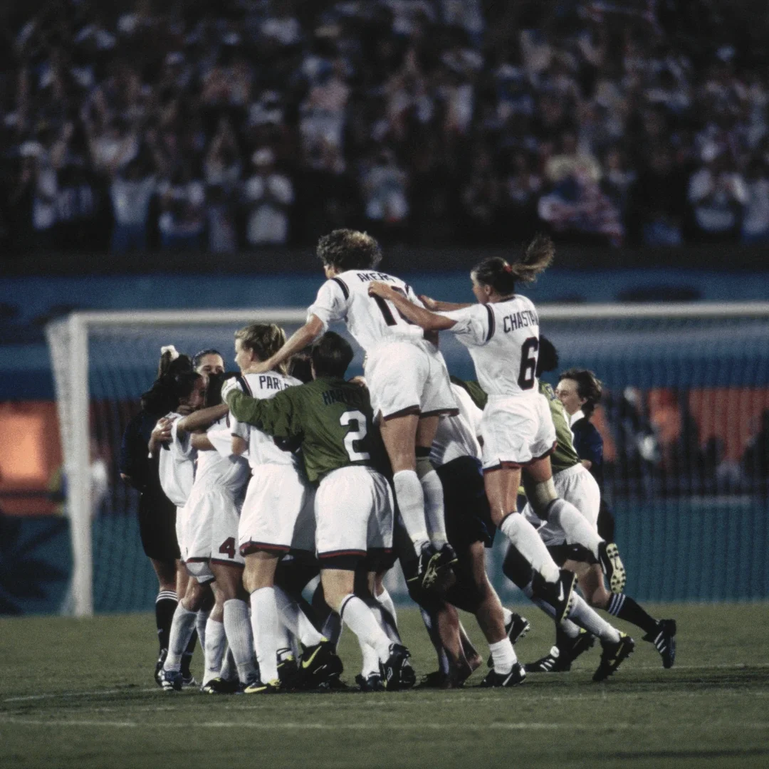 Integrantes de la selección nacional de Fútbol femenino de Estados Unidos, vistiendo camisetas blancas, pantalones cortos blancos y tacos, saltando en el aire en un abrazo grupal frente a una portería y el césped en los Juegos de Verano de Atlanta 1996.