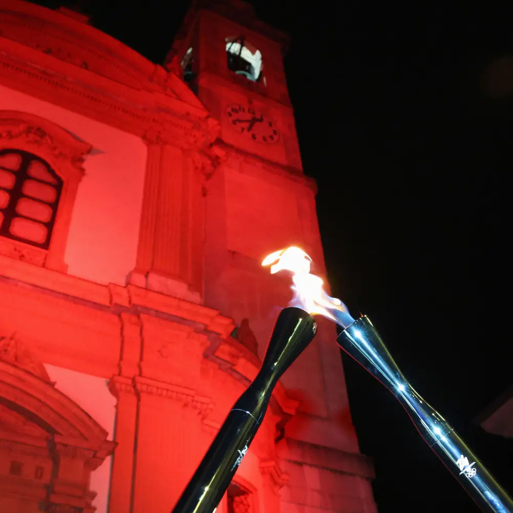 Two Games Torches touch each other in a criss-cross position with a clock tower in the background illuminated in red lighting. 