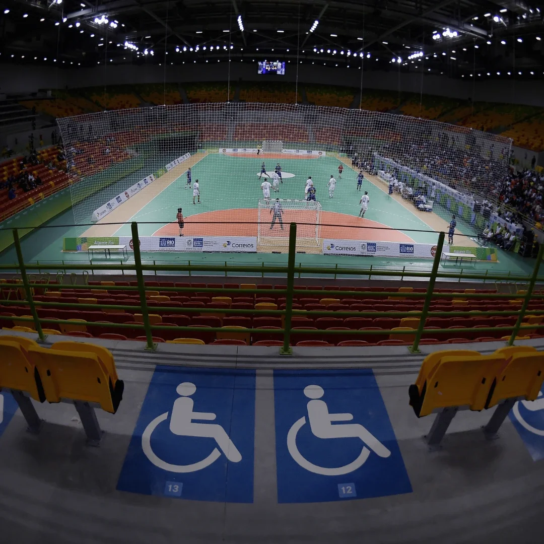 Image inside an arena where handball is being played. A row of wheelchair accessibility seating with the numbers 13 and 14 are in the foreground.
