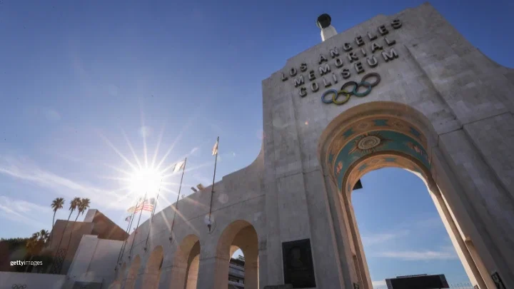 La entrada del Memorial Coliseum de Los Ángeles está fotografiada de frente durante el día. En la parte izquierda de la fotografía ondean banderas, incluida la estadounidense, y el Coliseum está flanqueado por palmeras. También hay una marca de Getty Images en la parte inferior izquierda de la foto.