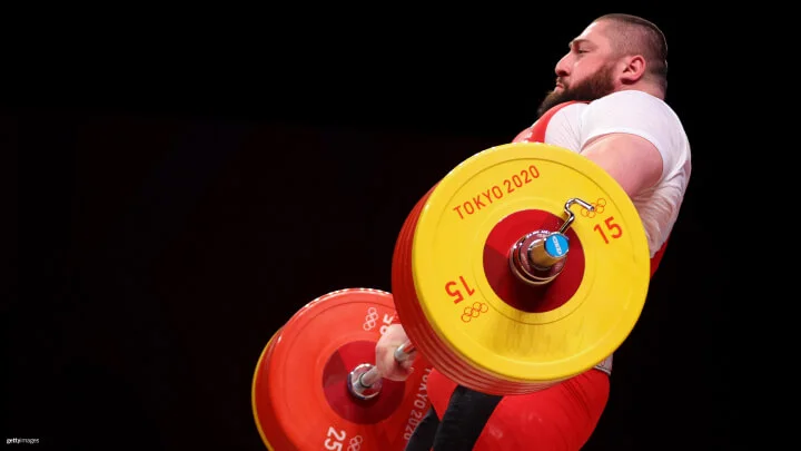 A weightlifter is performing a clean and jerk lift at the Tokyo 2020 Olympics. The athlete, wearing a white shirt and red singlet, is in the process of lifting a barbell with large red and yellow weight plates. The barbell has yellow and red plates with "Tokyo 2020" and the Olympic rings logo visible on them. The background is completely black.