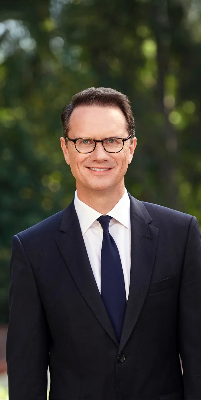 Photograph of Peter Rice in a suit with a white button down shirt and tie. Peter is standing in front of a bush or trees in the background. He is wearing glasses.