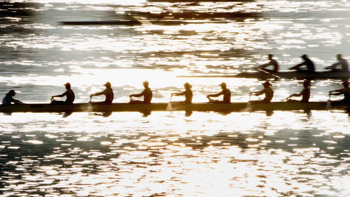 A rowing team is captured in silhouette, rowing in a long, narrow boat on a shimmering body of water. The sunlight reflects off the water, creating a sparkling effect. Each rower is synchronized, with their oars dipped into the water. Another rowing team is visible in the background.