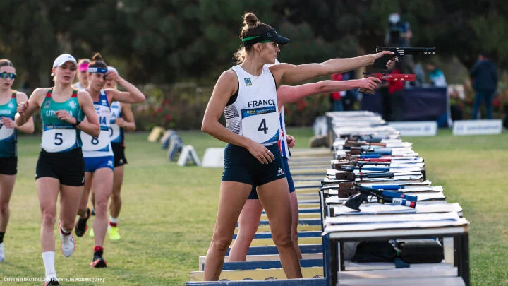 A female athlete from France competes in the shooting portion of a modern pentathlon, aiming a laser pistol while standing at a shooting station. Other competitors from different countries are seen running in the background, participating in the multi-discipline event. The scene takes place outdoors on a grassy field with various equipment laid out on tables.