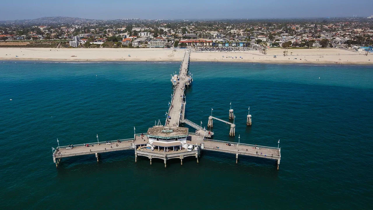 Fotografía aérea del Belmont Shore Pier en Long Beach, con el muelle extendiéndose recto hacia el océano y luego curvándose hacia la derecha e izquierda, donde al final se encuentra un pequeño edificio. Se pueden ver varias personas caminando por el muelle y en el fondo se observa una playa con arena, casas y edificios y un cielo azul.