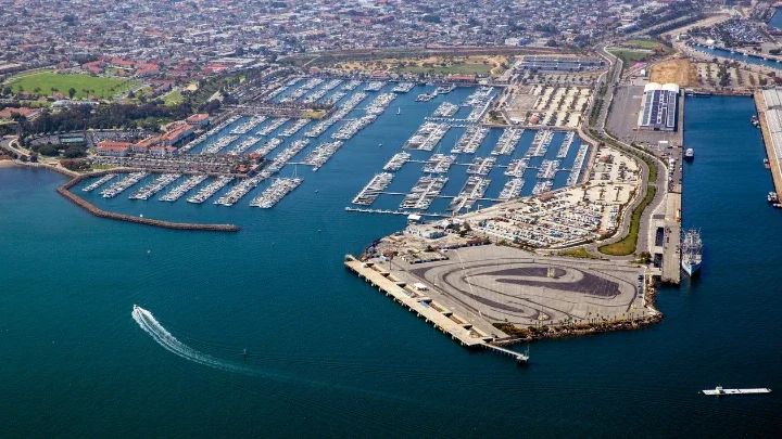 An aerial photograph of the Port of Los Angeles showing blue oceanfront and a marina full of boats with a helicopter landing pad in the foreground and in the background is a city scape featuring homes and buildings