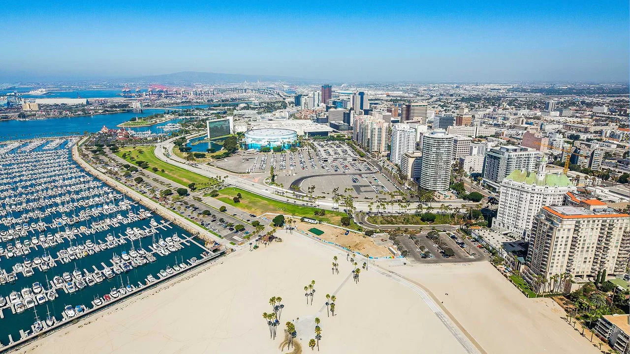 Aerial photograph of the watefront in Long Beach with a marina full of boats on the left, a sandy beach area in the front center, a parking lot in the back center, and numerous buildings and skyrises on the right, and a blue sky and city views in the background.