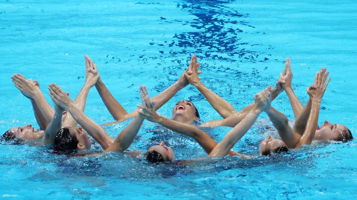 Se captura a un grupo de nadadoras artísticas realizando una rutina en una piscina azul brillante. Se encuentran en formación circular con las cabezas fuera del agua y los brazos extendidos hacia arriba, uniéndose las puntas de los dedos para crear una estrella. Las nadadoras muestran sonrisas y una expresión concentrada mientras llevan a cabo su coreografía.