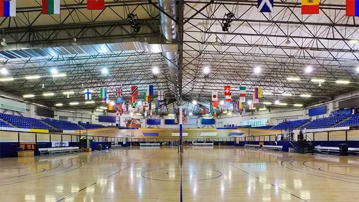 Photograph of an indoor velodrome with a brown wooden circular course and blue seating. There are numerous country flags hanging from the ceiling and a center court area with blue circles and lines drawn on a wooden floor.