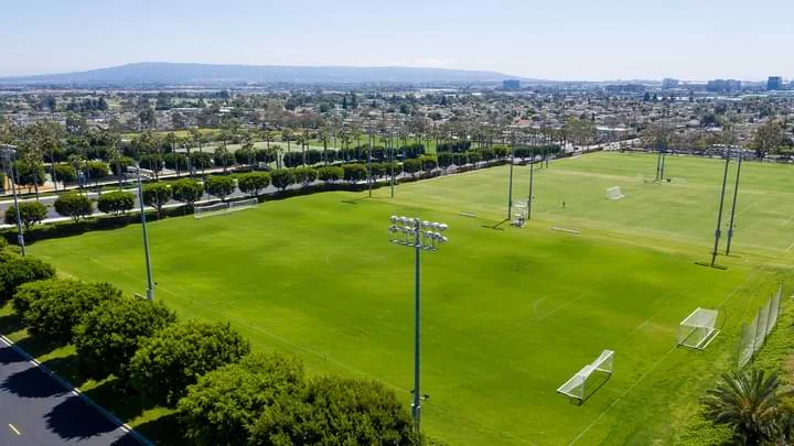 Photo from above of a large soccer field with goal boxes at each end and bright green grass, surrounded by green bushy trees as well as stadium lights. In the background is a city scape showing homes and buildings, blue sky and mountains in the distance.