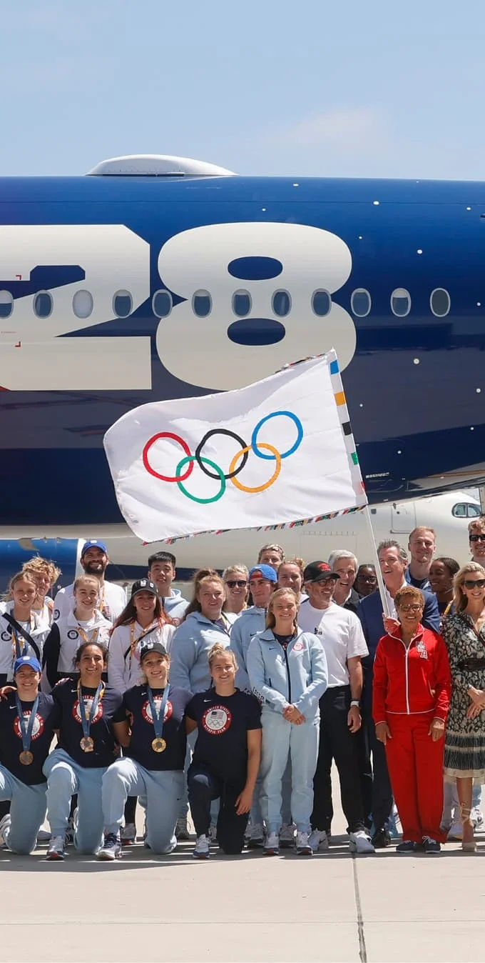 Fotografía de atletas del Team USA, funcionarios electos locales y ejecutivos de LA28 con la alcaldesa de Los Ángeles, Karen Bass, reunidos mientras el gobernador de California, Gavin Newsom, sostiene la bandera olímpica frente al avión Delta Olympic Flag Flight. 