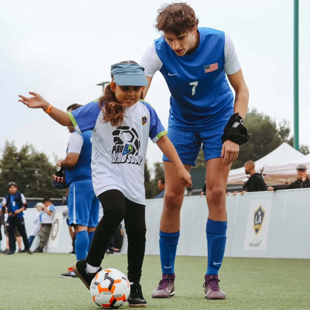 A young girl is kicking a soccer ball while a coach stands behind her supporting her